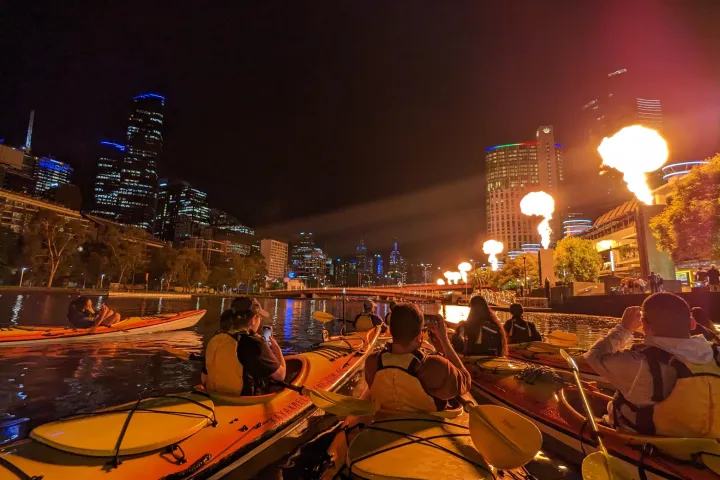 Group of kayakers on a river at night with city lights and fire installations.
