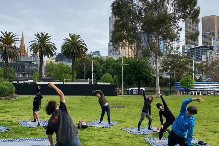 People doing yoga on mats in a city park with trees and skyscrapers.