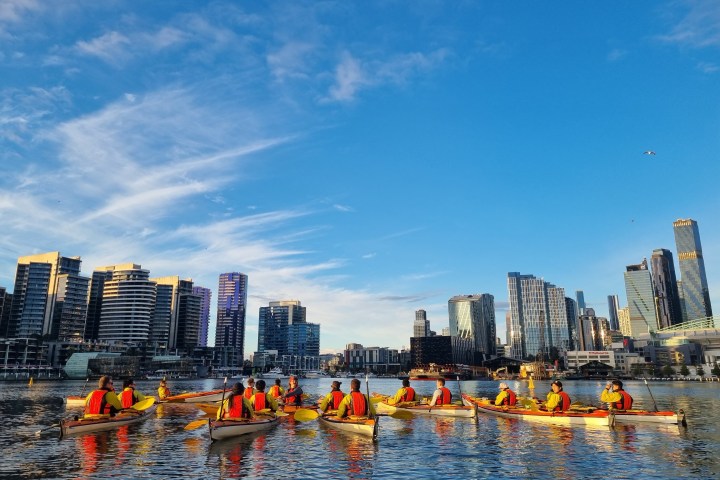 Group kayaking on a river against a cityscape backdrop under a blue sky.
