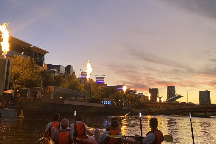 People in kayaks on a river at sunset with city skyline in background.