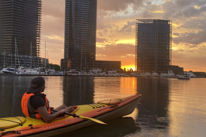 Person kayaking on calm water at sunset, city skyline in background.
