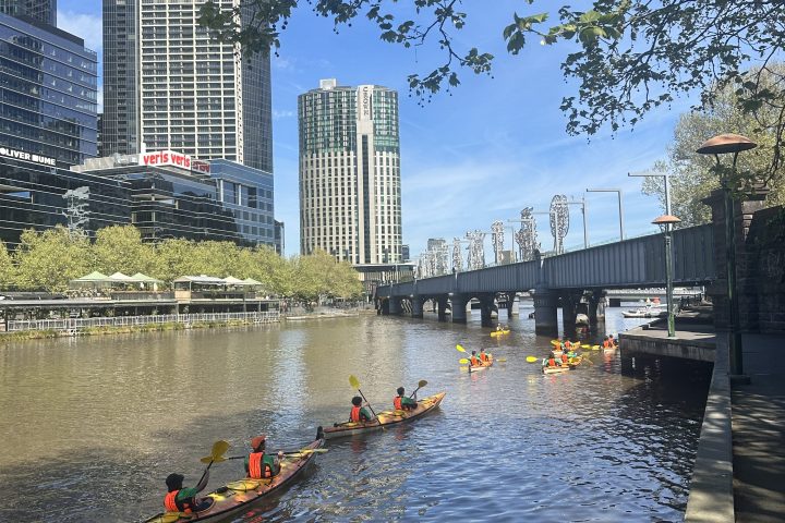 Kayakers paddle on a river near tall city buildings under a clear blue sky.