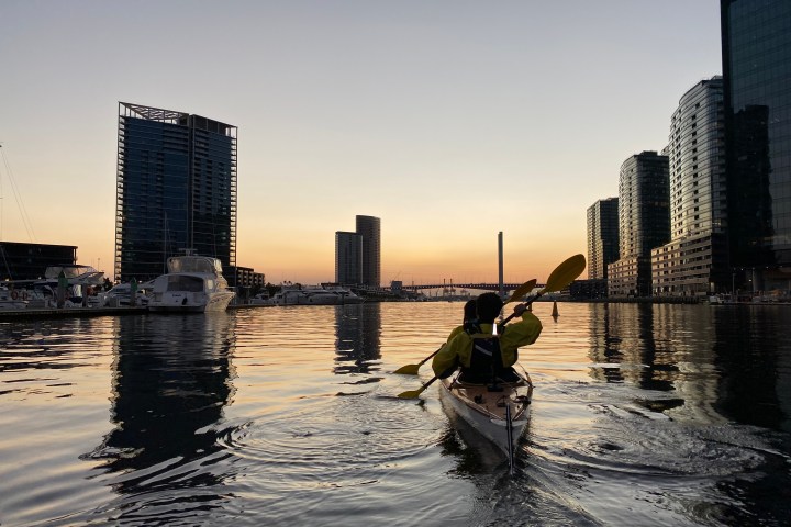 Person kayaking on calm water at sunset, surrounded by modern city buildings.