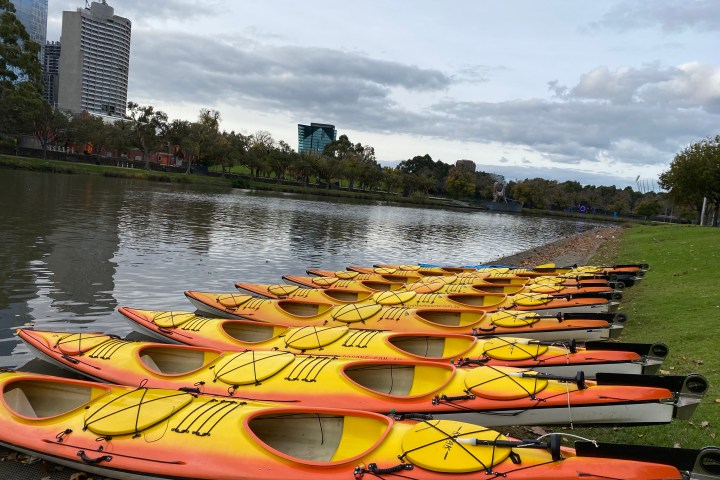 Rows of yellow kayaks on a riverbank with city buildings in the background.